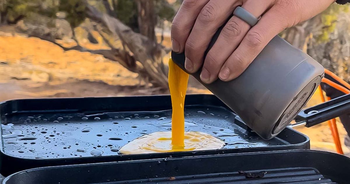 pouring mixed eggs from mug onto outdoor grill at campsite